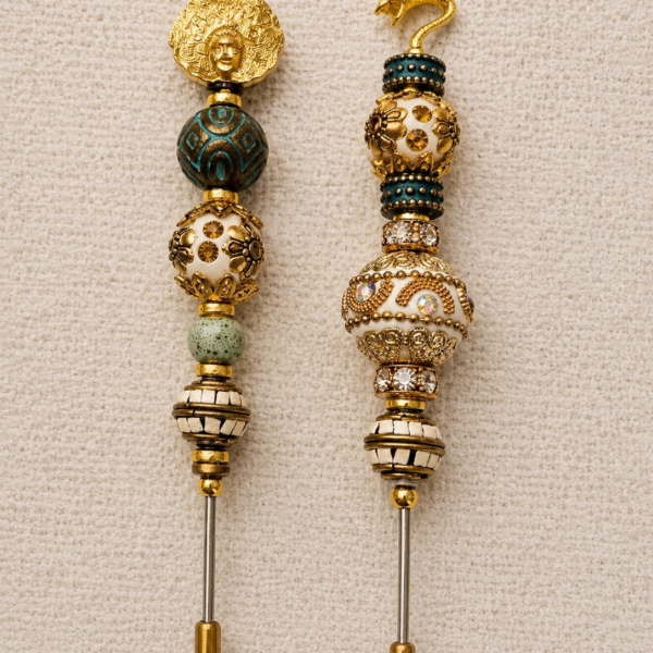 Pair of ivory, teal, and gold beaded cigar nubbers with a medallion face topper on one piece and a serpent topper on the other, displayed on a soft beige background.