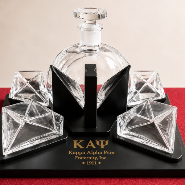 Faceted glass decanter set with four angular tumblers on a black Kappa Alpha Psi display stand against a deep red surface.